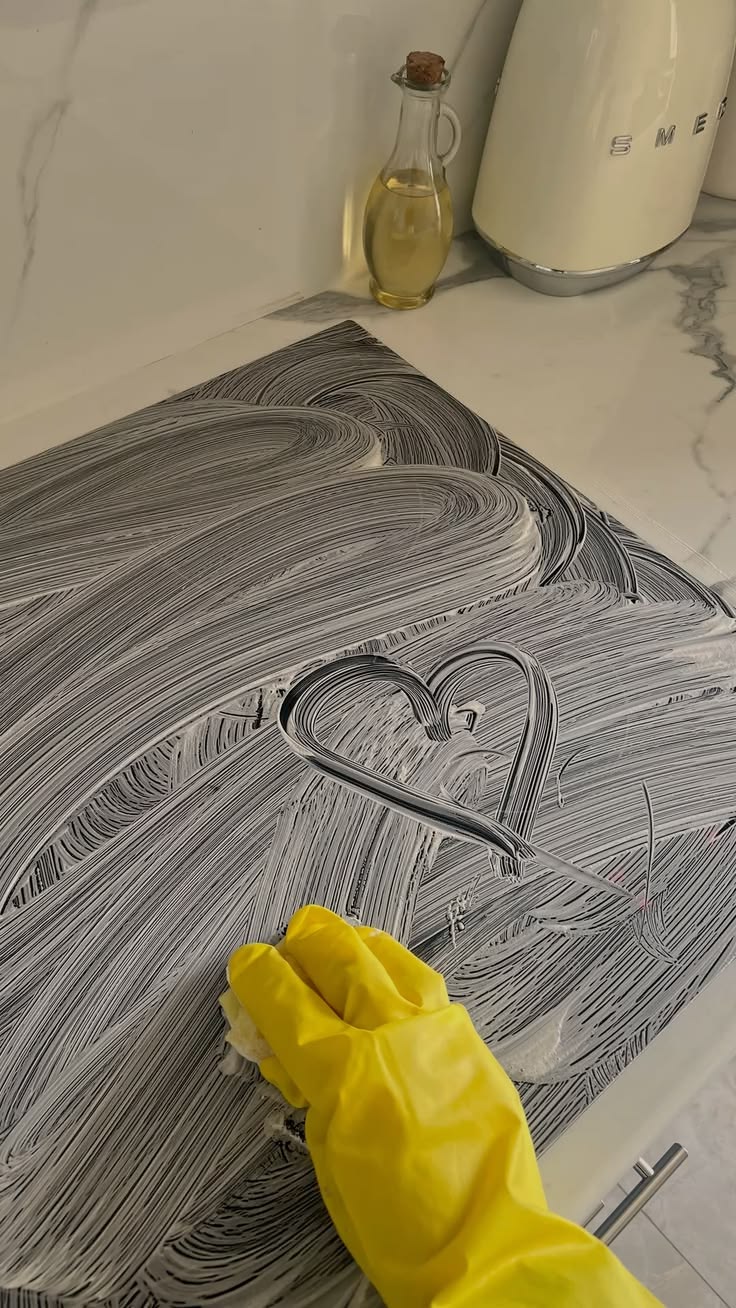 A gloved hand wiping cleaning foam across a dark stovetop, with a heart drawn in the suds